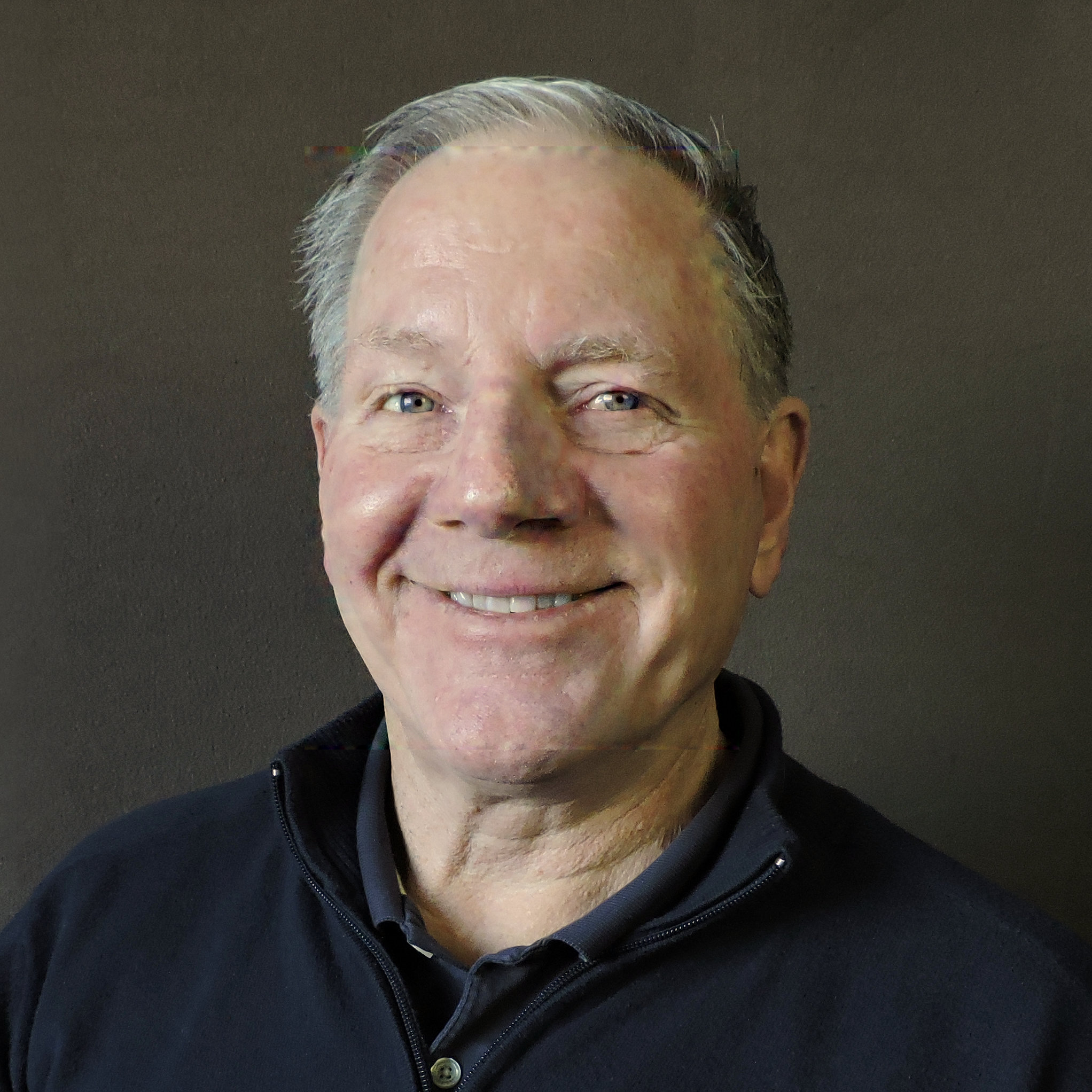 Pat Runnion in a blue zip up shirt smiling in front of a grey background